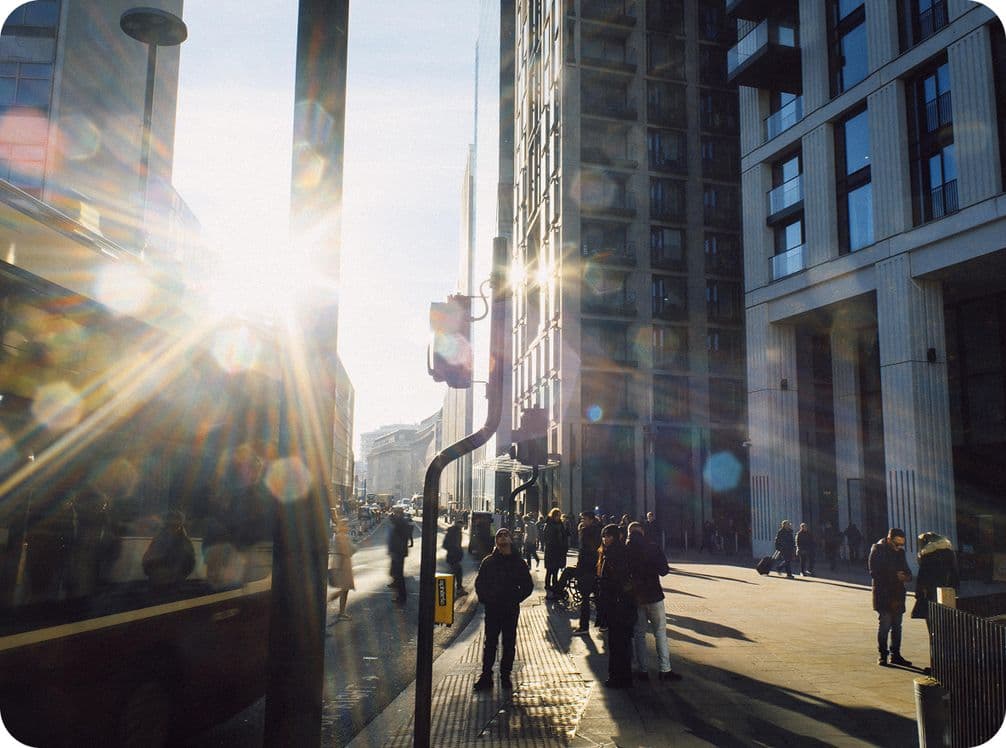City street scene with people walking, sunlight creating lens flares, and tall buildings casting long shadows.
