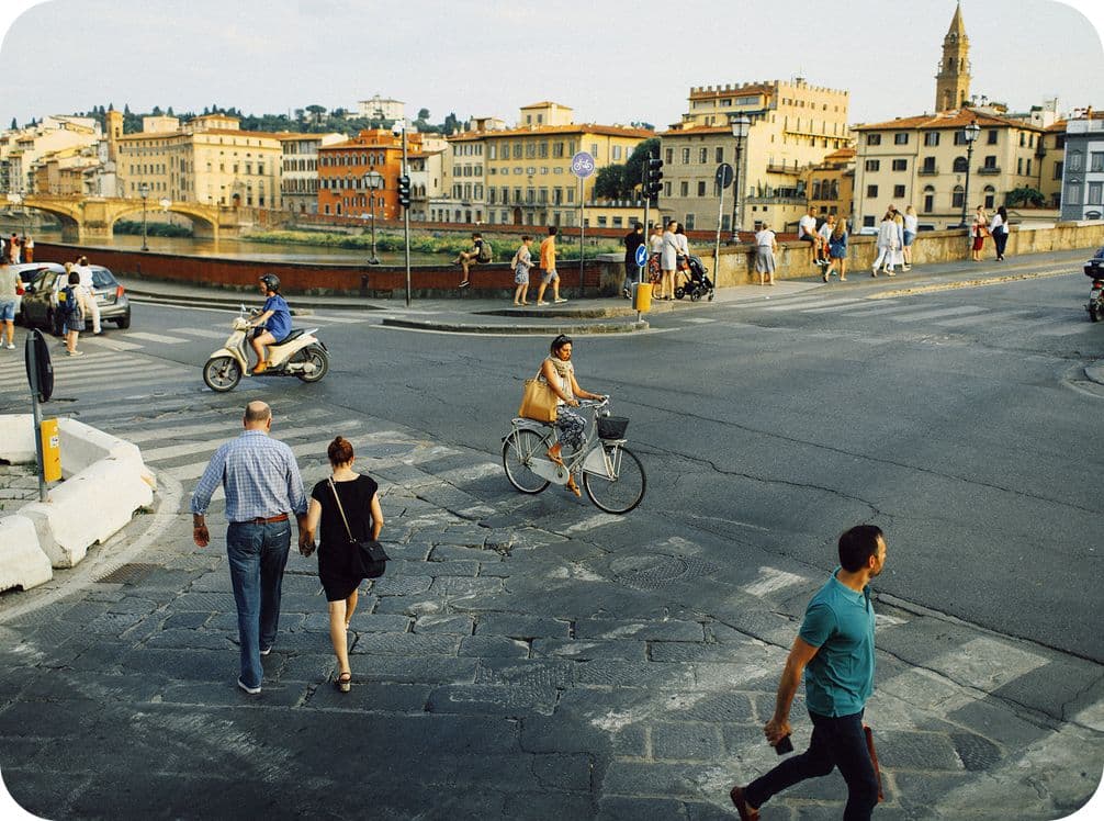 People walking and cycling near a river in a European city, with historic buildings and a church tower in the background.