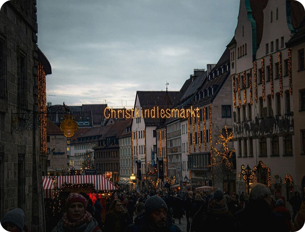 A bustling Christmas market with festive lights, crowded with people, and a "Christkindlesmarkt" sign overhead in a European city street.