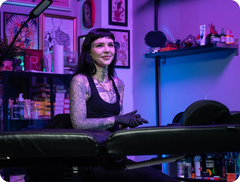 Tattoo artist with black gloves sits in a studio with purple lighting, surrounded by art and equipment.