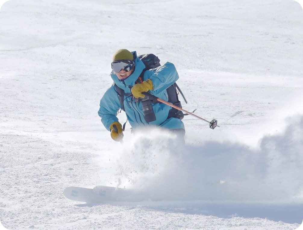 A skier in a blue jacket and green hat glides through fresh snow, creating a spray, under a clear sky.