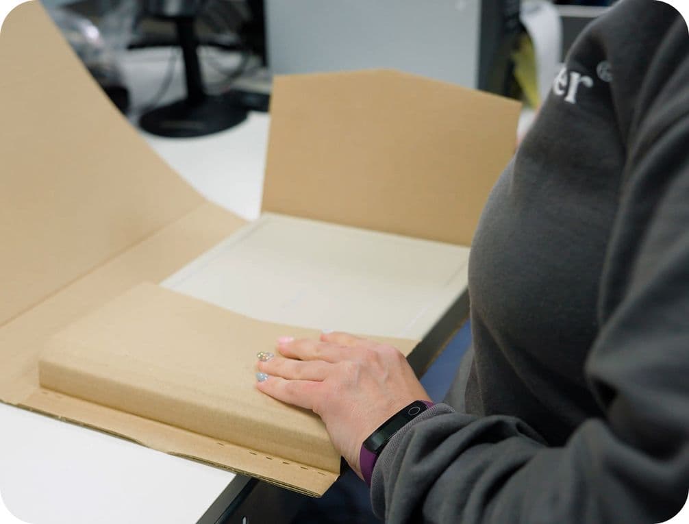 Person packaging a flat item in a cardboard envelope on a desk, wearing a dark sweater and a fitness tracker.