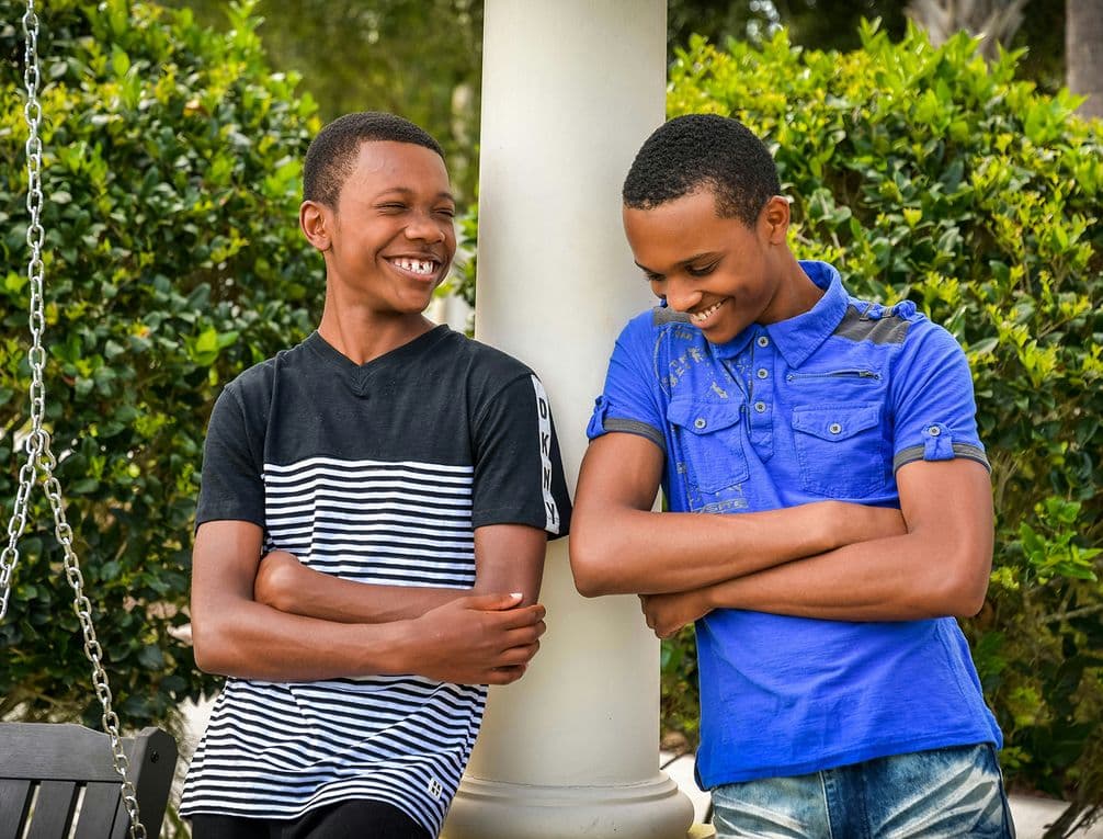 Two teenage boys leaning against a white column outdoors, smiling and laughing with arms crossed, greenery behind them.