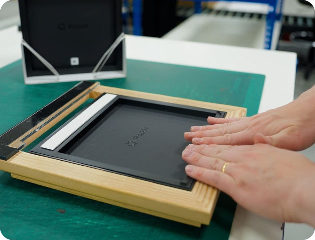 Hands assembling a wooden picture frame on a table, with another frame in the background.