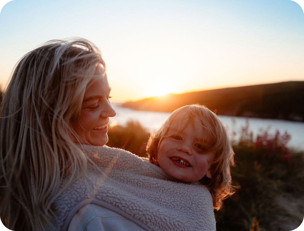 A woman with long hair holds a smiling child wrapped in a blanket, with a sunset over a river in the background.