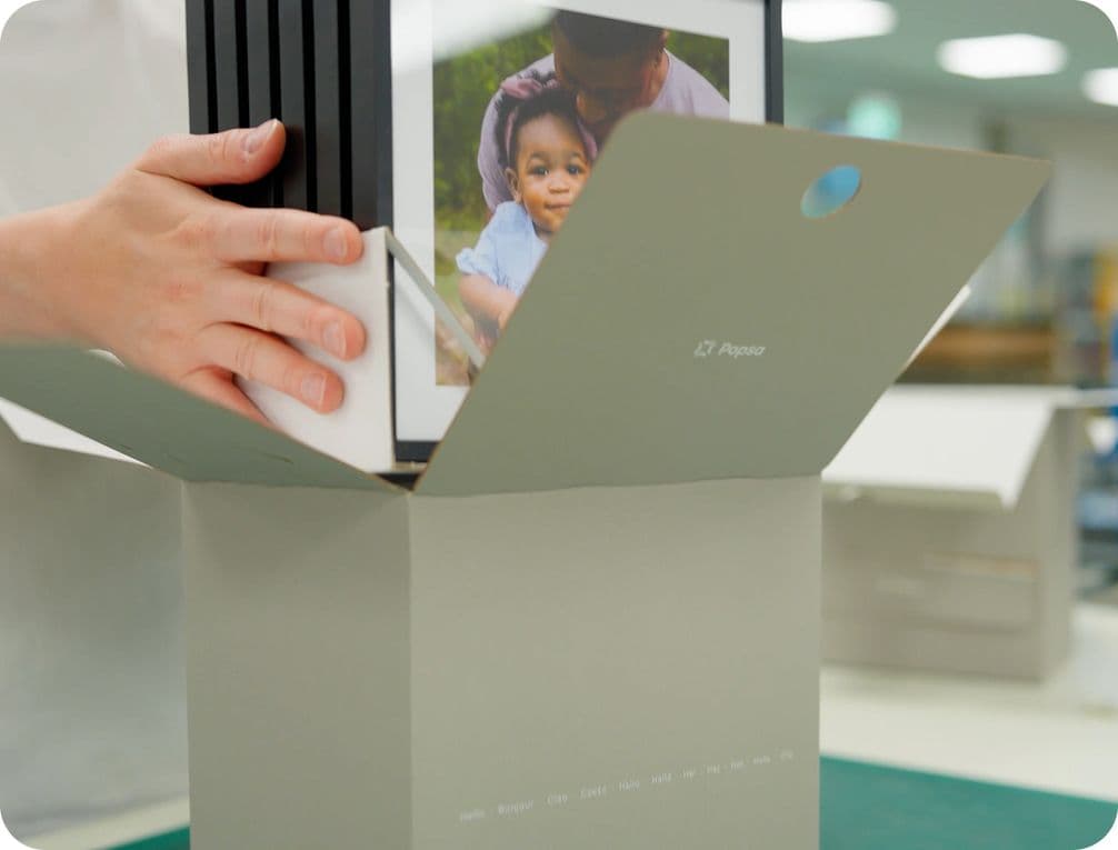Hands placing a stack of framed photos, with a picture of a child, into a gray box labeled "G7 Paper" in a well-lit room.