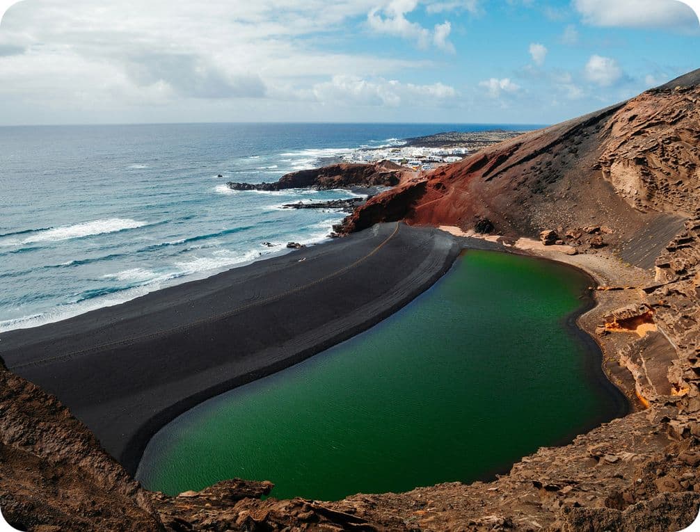 A vivid green lagoon bordered by black sand and rocky cliffs, adjacent to the ocean under a partly cloudy sky.