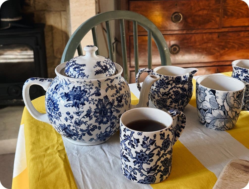 Blue floral teapot and cups on a yellow and white tablecloth, with a wooden chair and cabinet in the background.