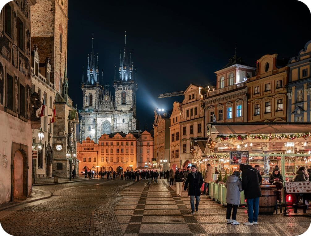 A bustling Prague Christmas market at night with a lit cathedral, historic buildings, and people enjoying a festive market.