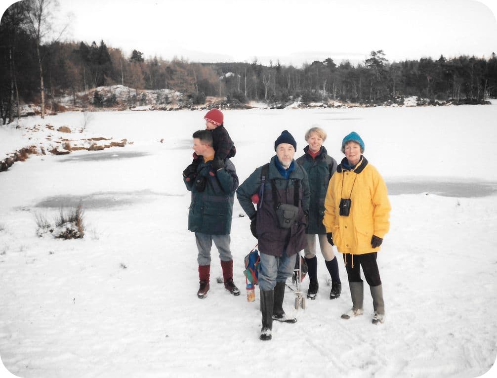 Five people in winter clothing stand on a snowy landscape near a frozen lake, surrounded by trees. One person carries a child.