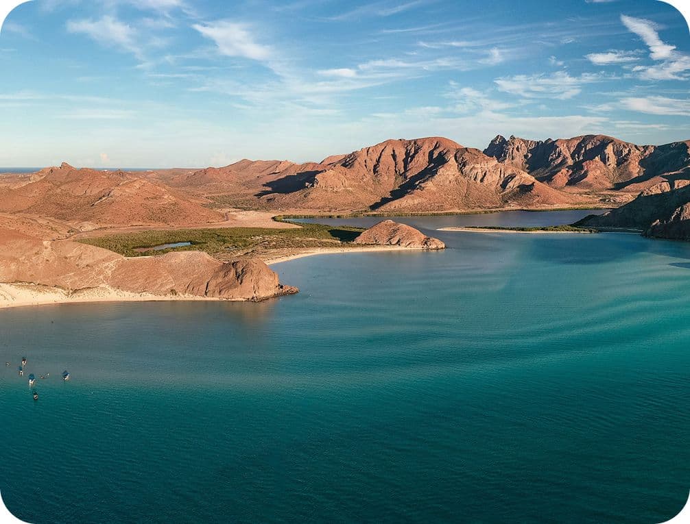 Aerial view of a serene bay with turquoise water, surrounded by rugged, reddish-brown mountains under a clear blue sky.