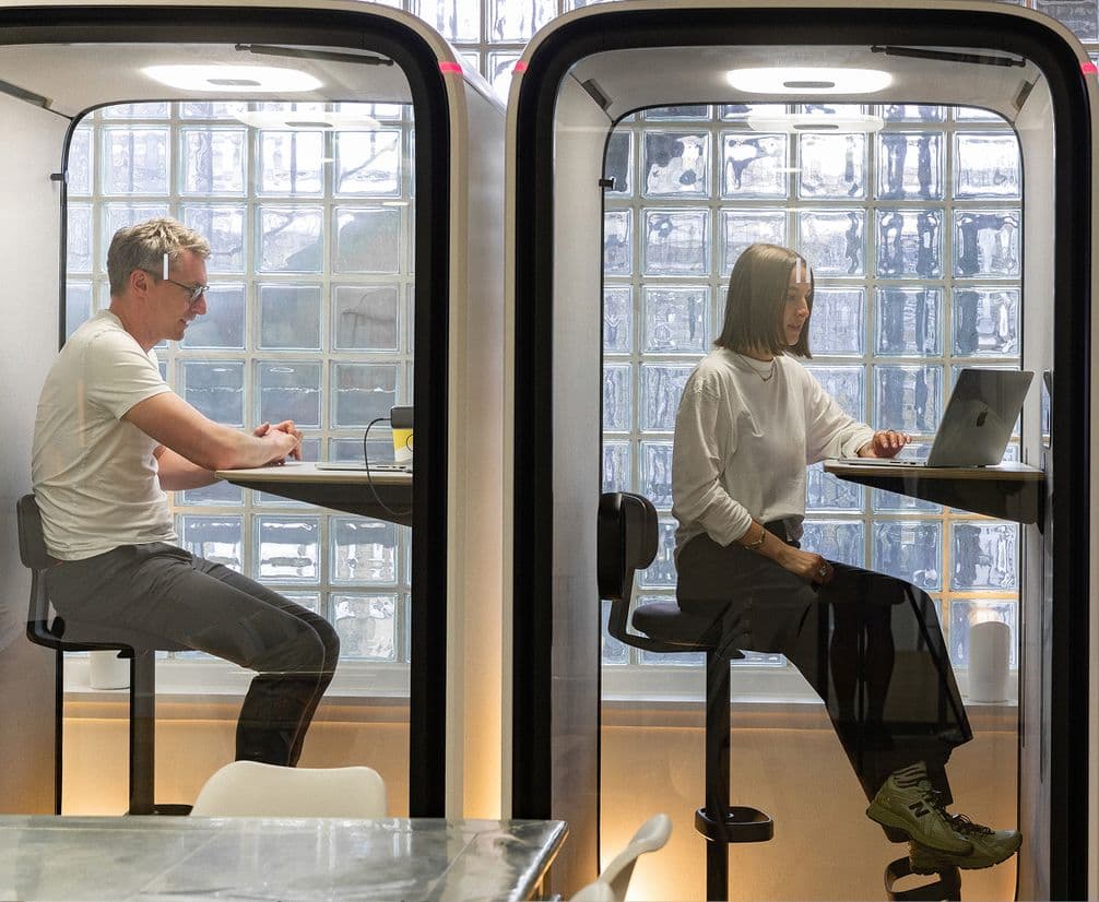 Two people working on laptops inside separate, transparent office pods with curved glass walls and bar stools.