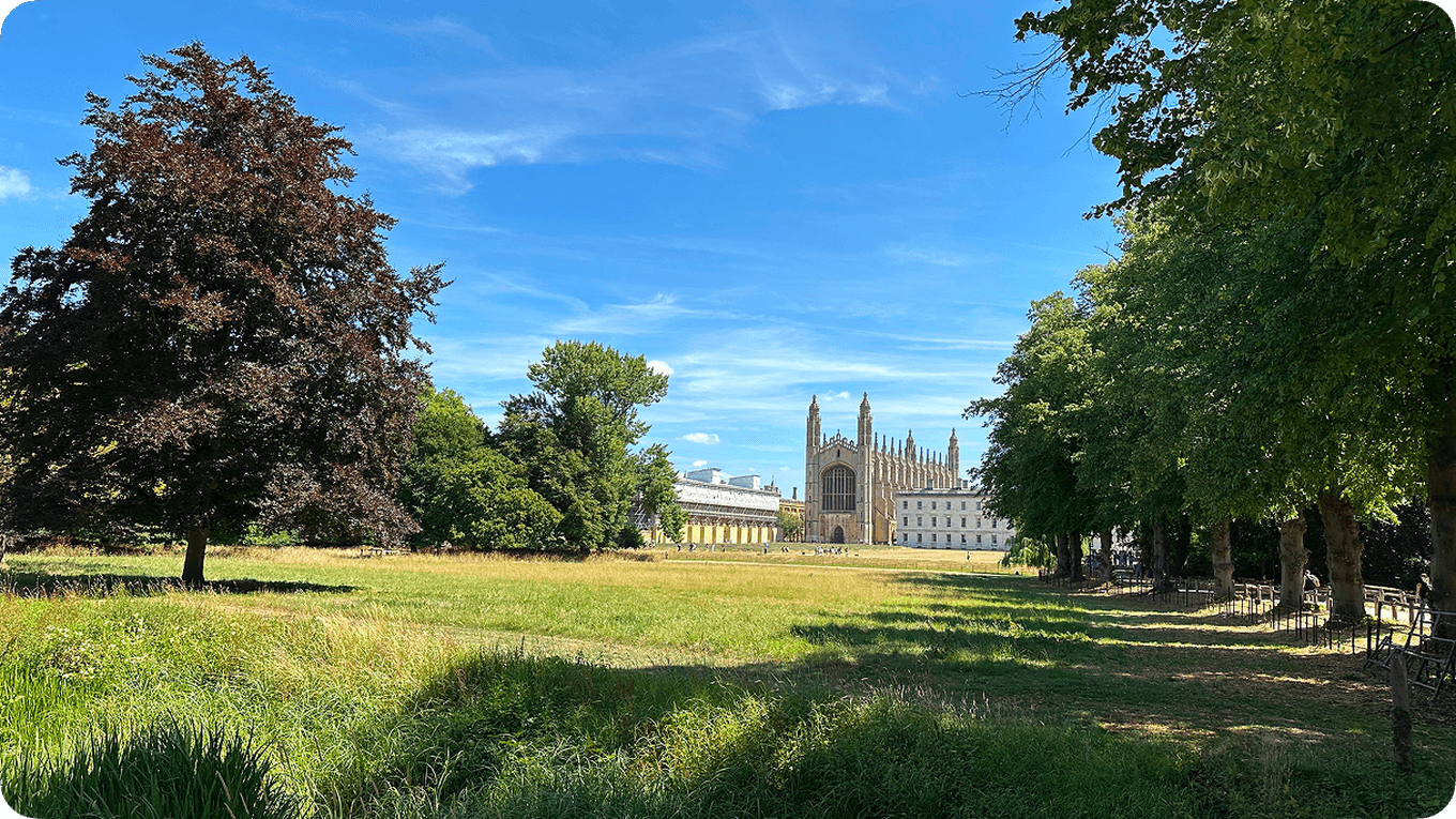 A scenic view of a grassy field with trees and the University of Cambridge under a clear blue sky.