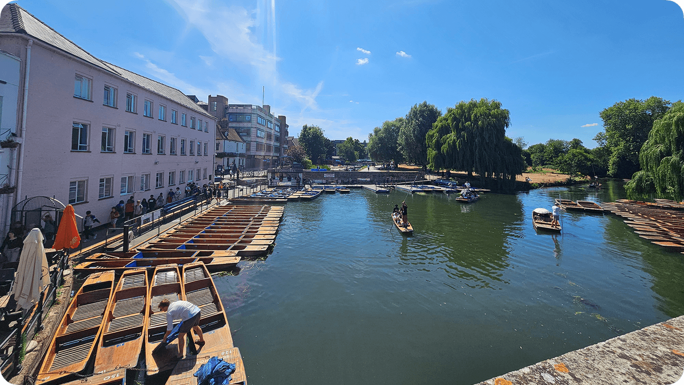People enjoying a sunny day on a river in Cambridge with wooden boats and lush greenery, surrounded by buildings.