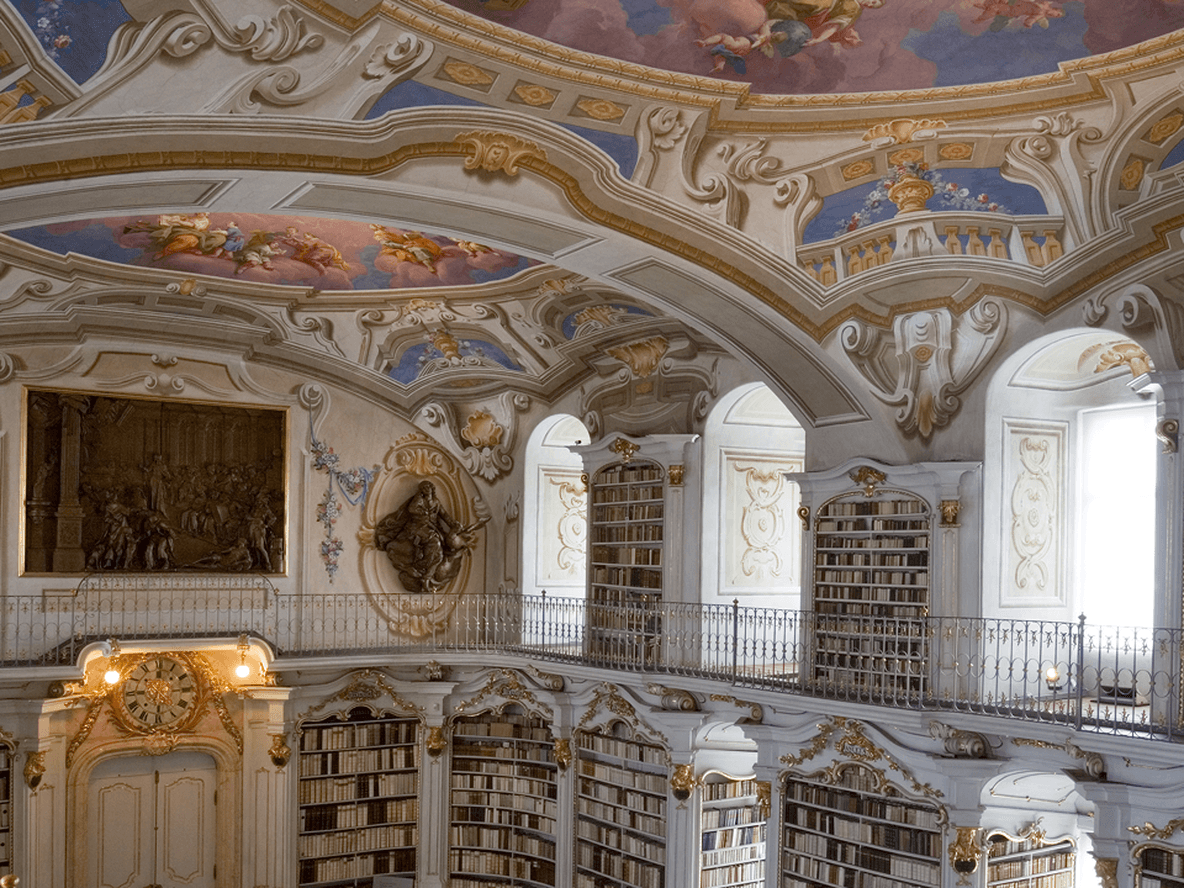 Ornate library interior with arched ceiling, elaborate frescoes, tall bookshelves, and decorative sculptures. Bright natural light illuminates the space.