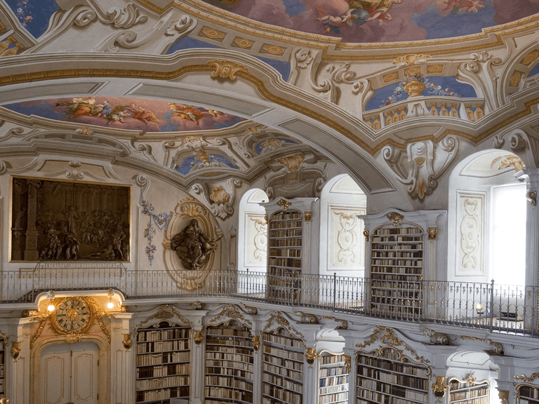 Ornate library interior with arched ceiling, elaborate frescoes, tall bookshelves, and decorative sculptures. Bright natural light illuminates the space.