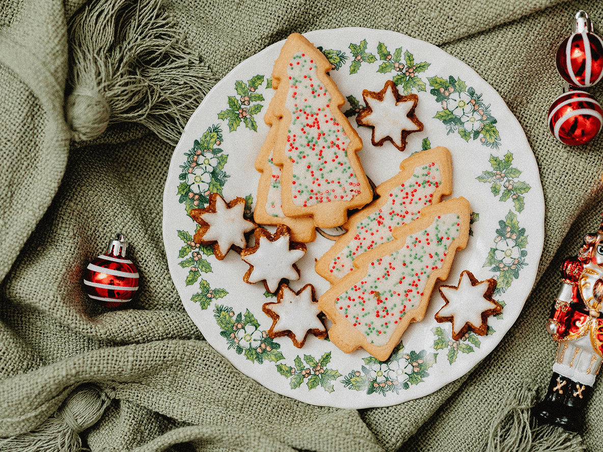 Plate of Christmas-themed cookies on a green cloth, surrounded by red ornaments and a nutcracker figurine.