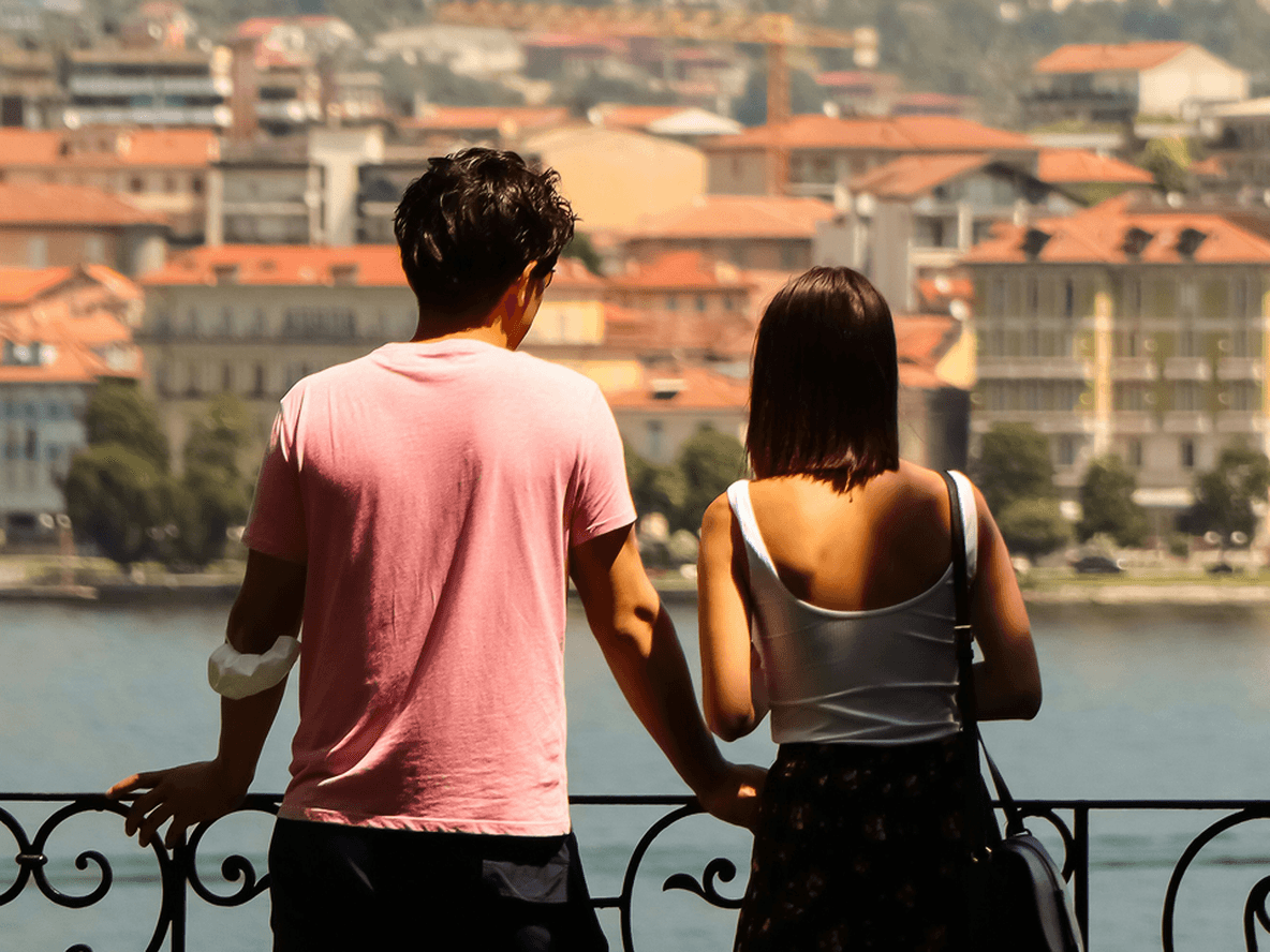 A couple leans on a railing, overlooking a scenic view of a lake and a town with red-roofed buildings in the background.