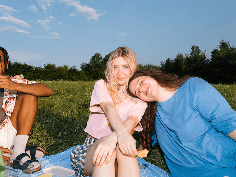 Three people enjoying a sunny picnic on a grassy field, with one person resting their head on another's shoulder, surrounded by trees.