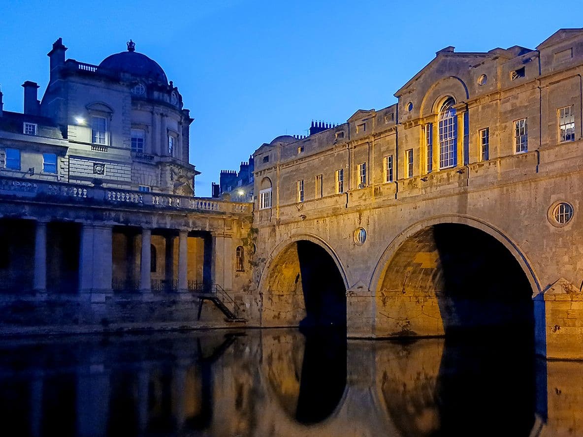 Illuminated historic stone bridge and buildings reflected in calm river water at twilight, under a clear blue sky.