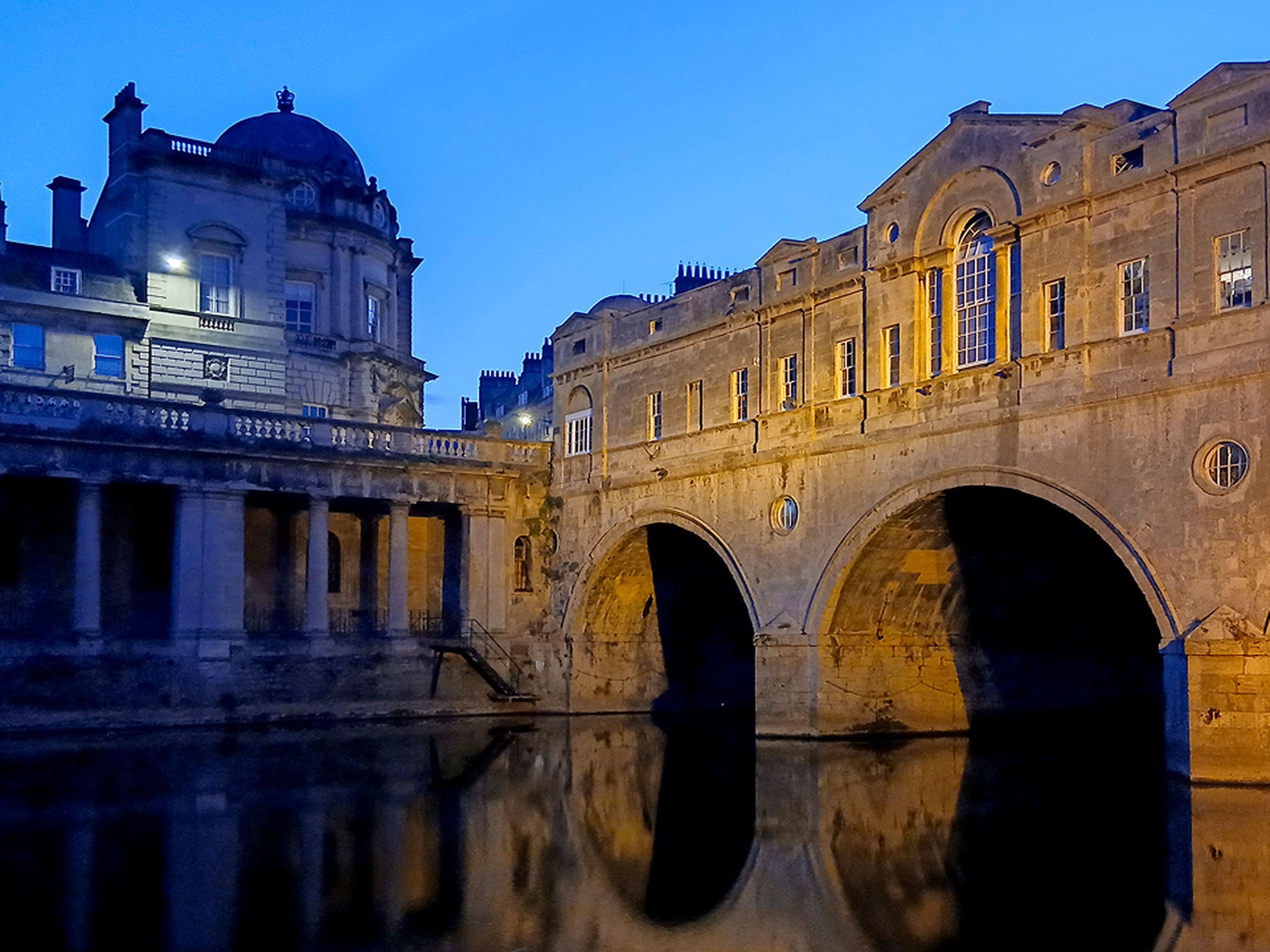 Illuminated historic stone bridge and buildings reflected in calm river water at twilight, under a clear blue sky.