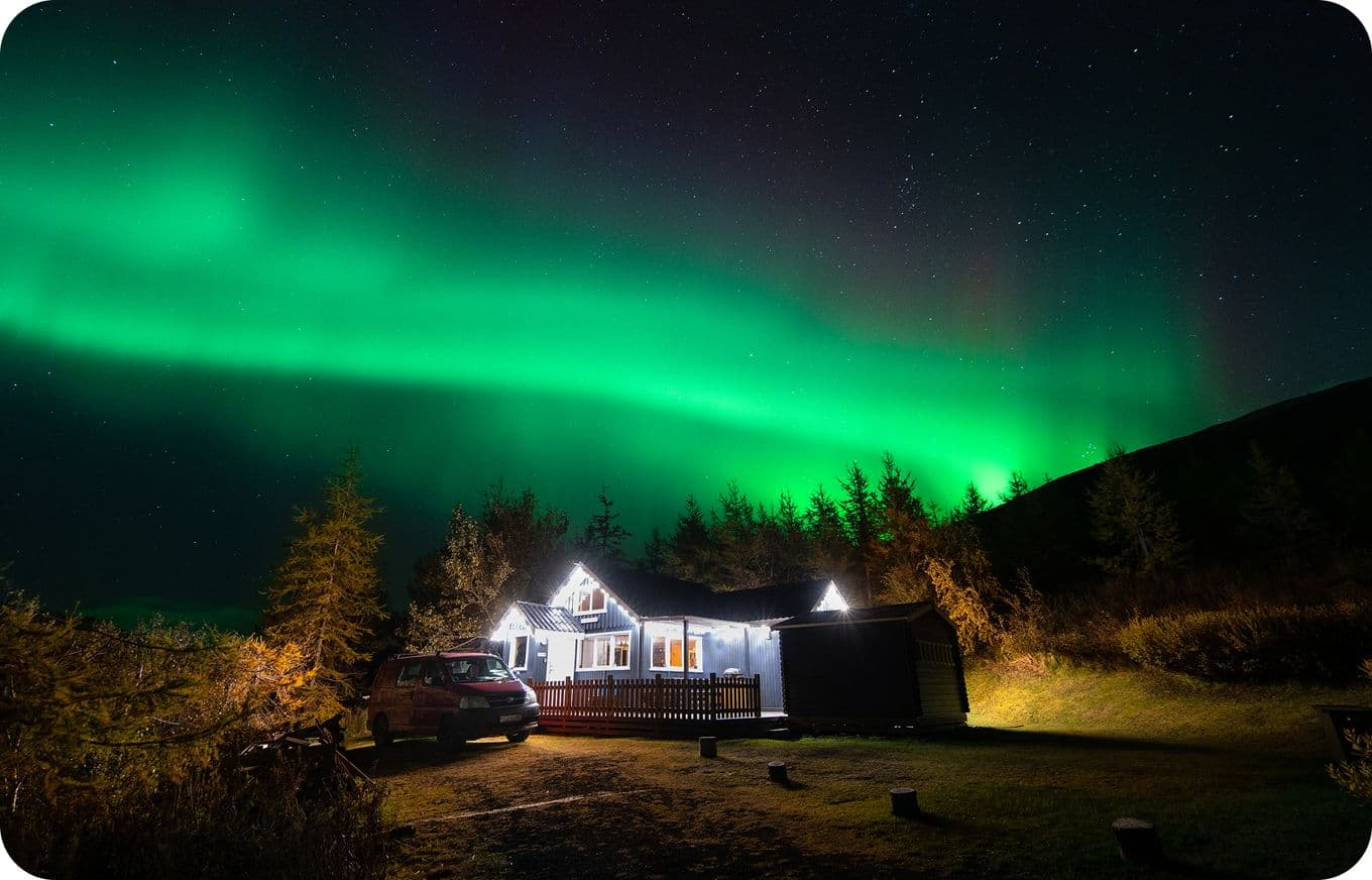 A cozy cabin with a parked car under a vibrant green aurora borealis in a starry night sky, surrounded by trees and a hill.