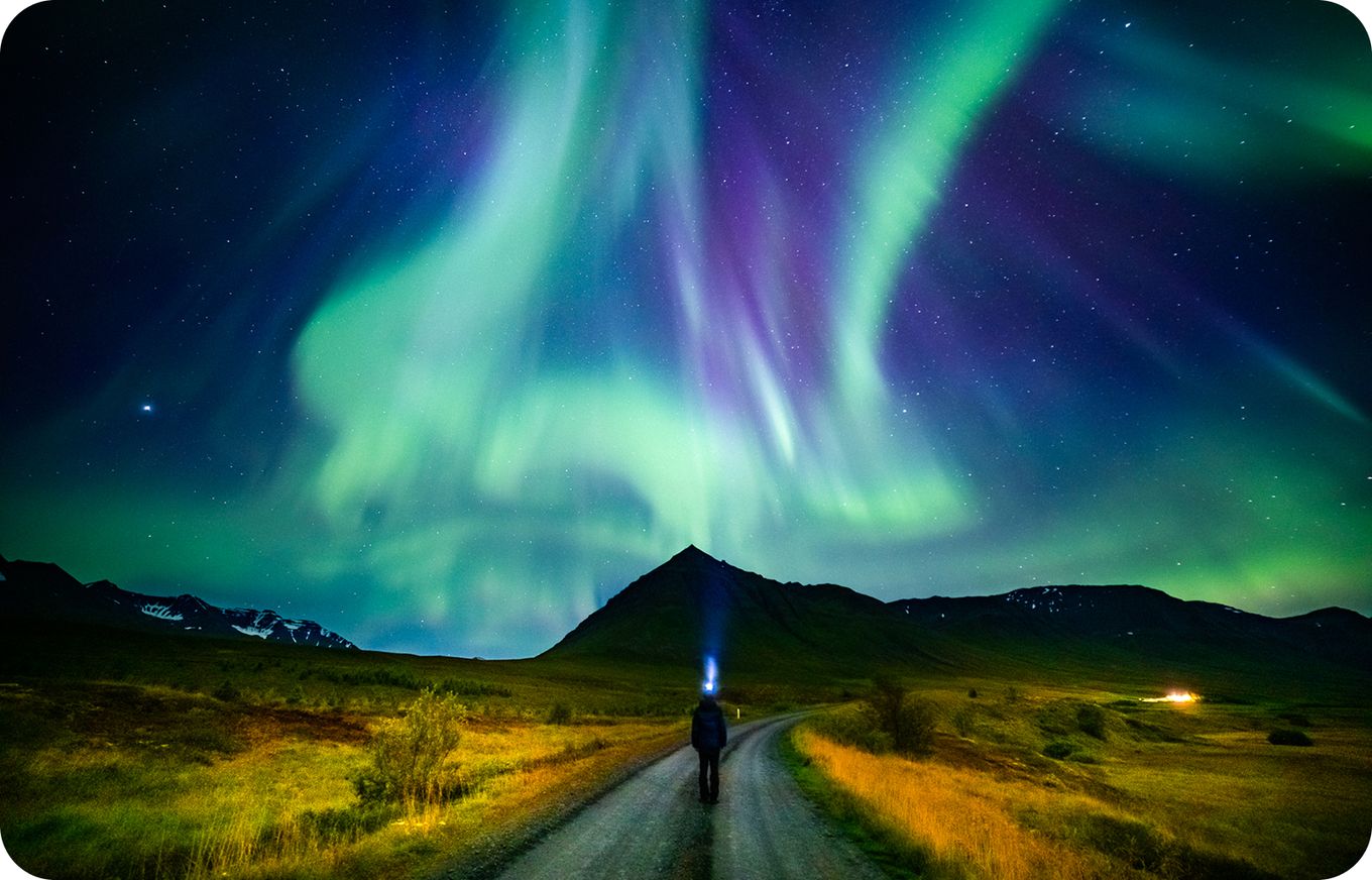 Person on a path under vibrant green and purple Northern Lights, with a mountain silhouette in the background.