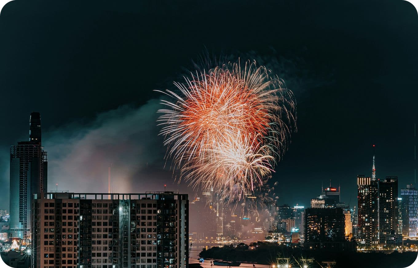Fireworks light up the night sky over a cityscape, with tall buildings illuminated and smoke trailing from the bursts.