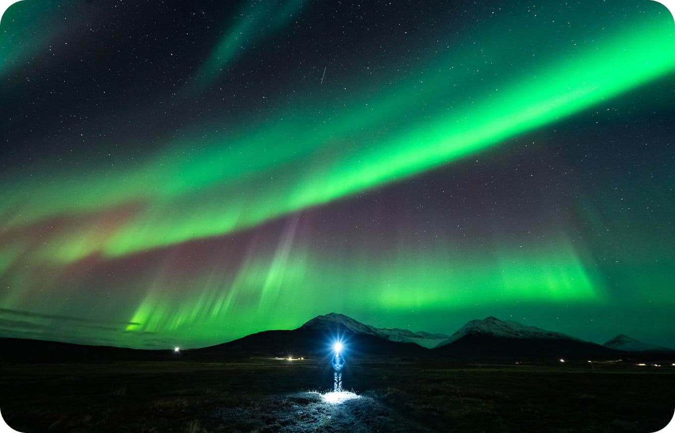 Person with a flashlight stands under vibrant green and purple Northern Lights in a starry sky over snow-capped mountains.