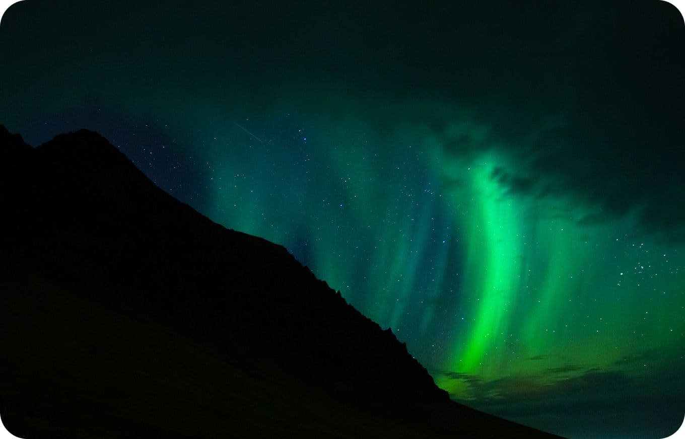 Northern lights with vibrant green and blue hues illuminate the night sky over a dark, silhouetted mountain landscape.