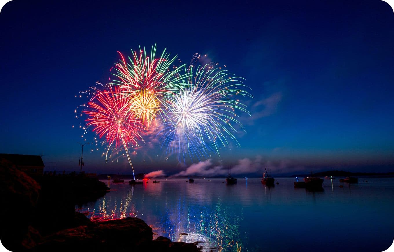 Colorful fireworks burst over a calm lake at dusk, reflecting vibrant reds and greens on the water, with silhouetted boats below.
