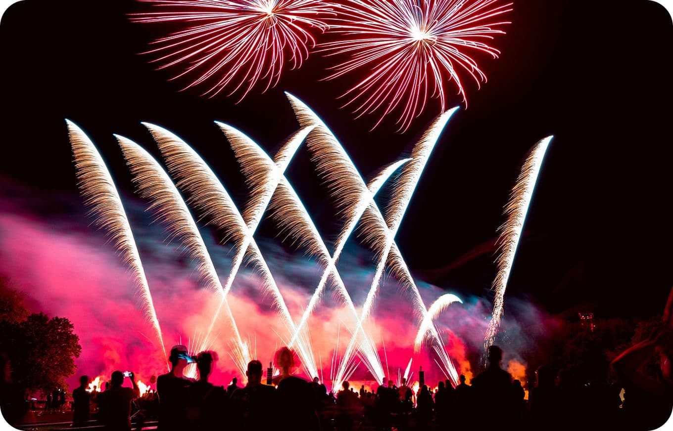 A crowd watches a vibrant fireworks display at night, featuring red and white bursts and streaks against a dark sky.