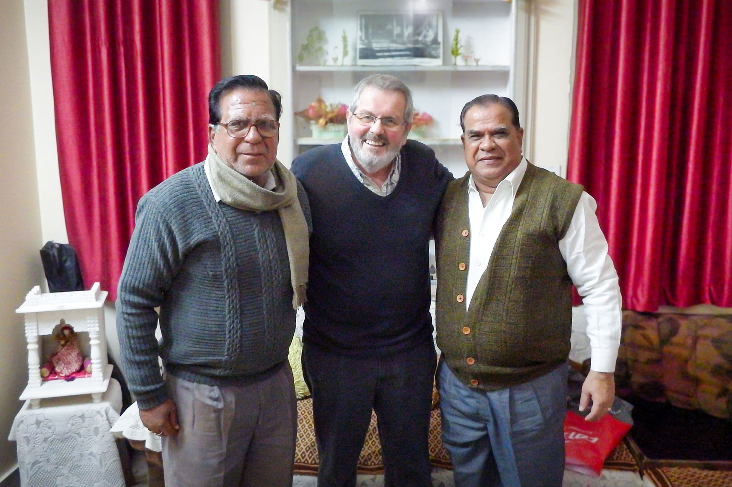 A smiling Geoffrey standing in a living room with his arms round Radheshyam and Satynarain Joshi on either side of him