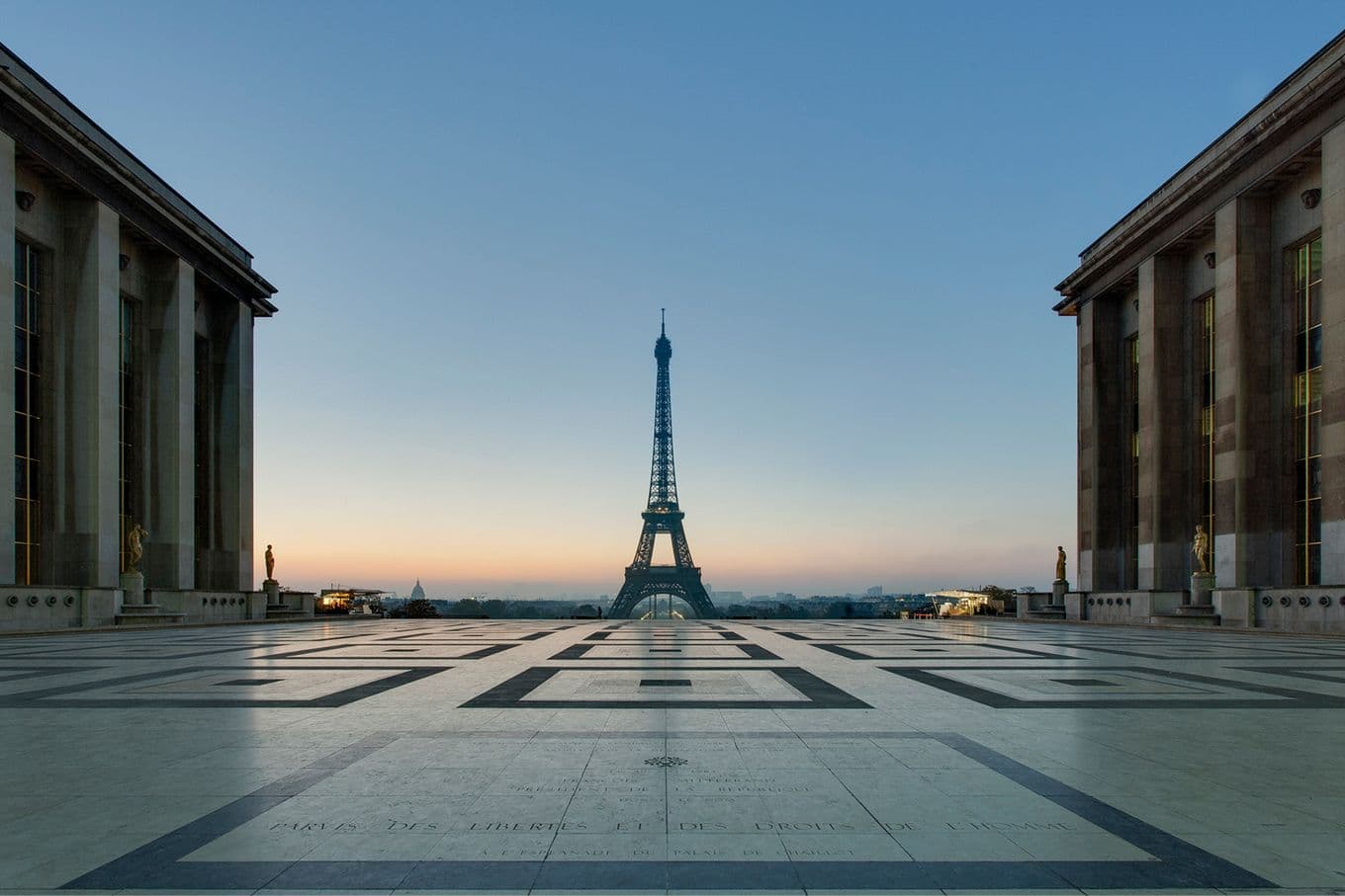 The Eiffel Tower viewed from Trocadéro at sunrise, framed by two symmetrical buildings and a patterned plaza in the foreground.