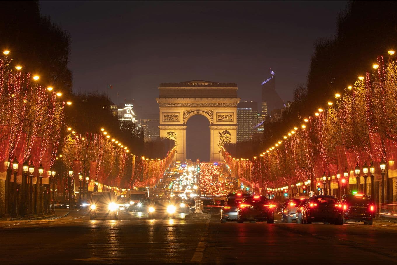 Champs-Élysées at night with festive lights, cars on the road, and the Arc de Triomphe illuminated in the background.