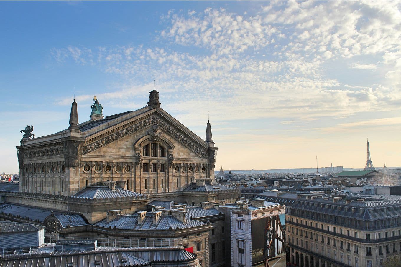 Aerial view of the Palais Garnier in Paris, with ornate architecture, under a partly cloudy sky. The Eiffel Tower is visible in the distance.