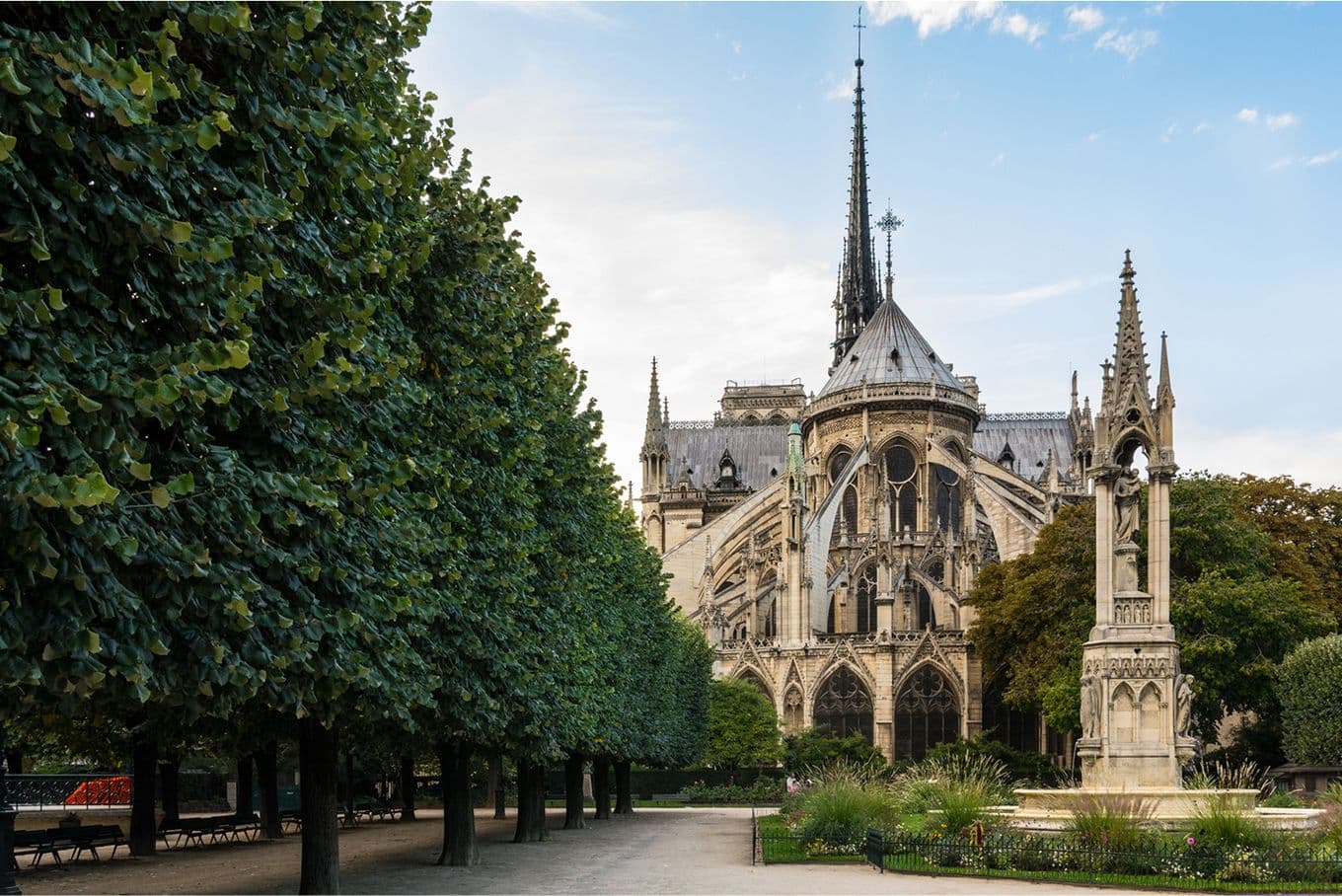 Rear view of Notre-Dame Cathedral with a garden path lined by trees and a statue in the foreground under a partly cloudy sky.