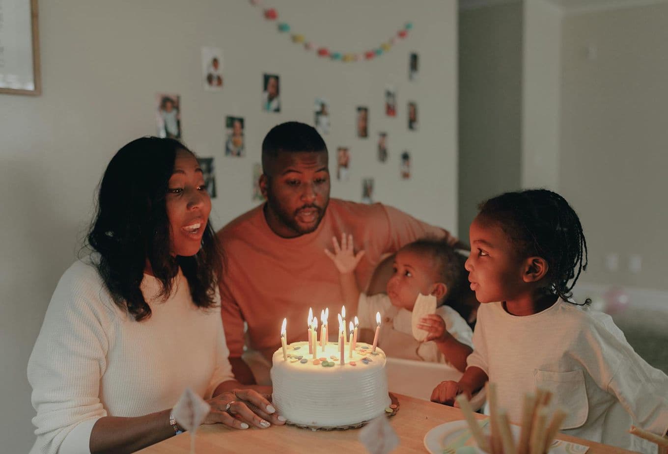 Family celebrating a birthday with a lit cake. Two children and two adults gather around the table in a warmly decorated room.