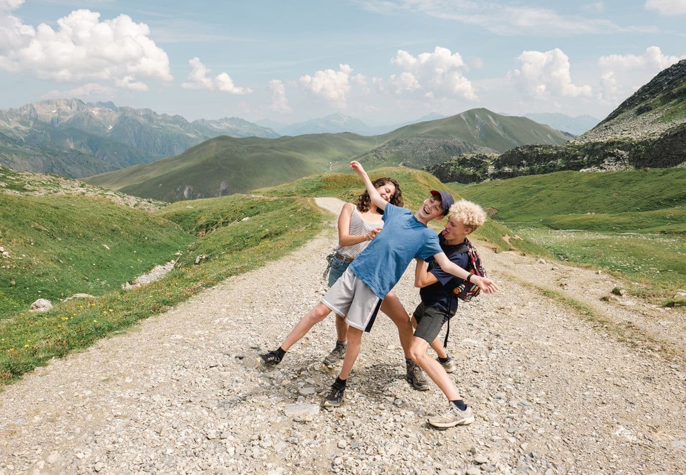 Three people joyfully pose on a rocky mountain path, surrounded by green hills and distant peaks under a blue sky with fluffy clouds.