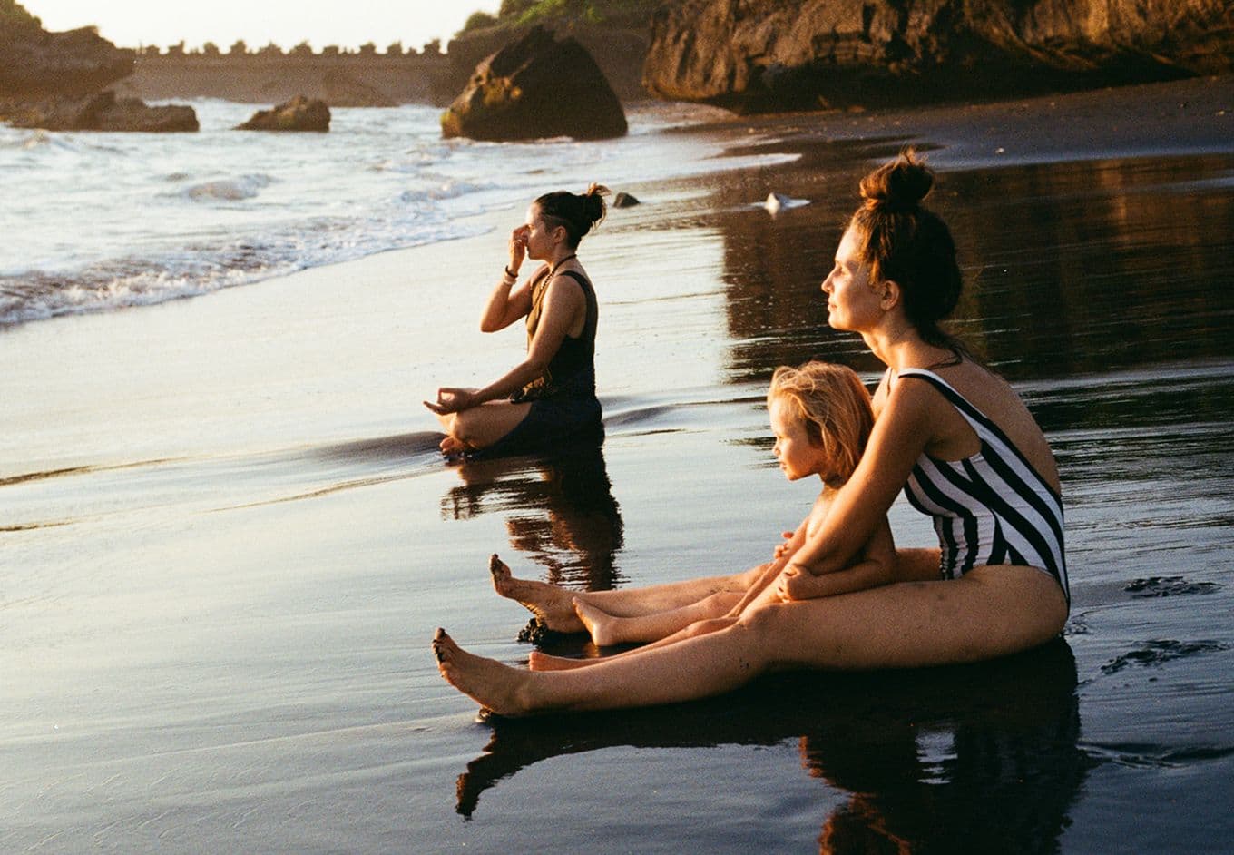 Two women and a child sat meditating at the water's edge on a beach.