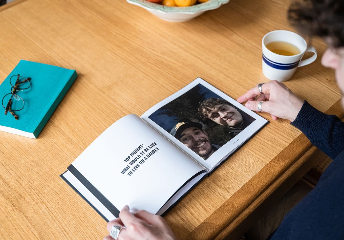 Person looking at an open photo book with a picture of two smiling individuals. A cup, glasses, and a teal notebook are on the wooden table.