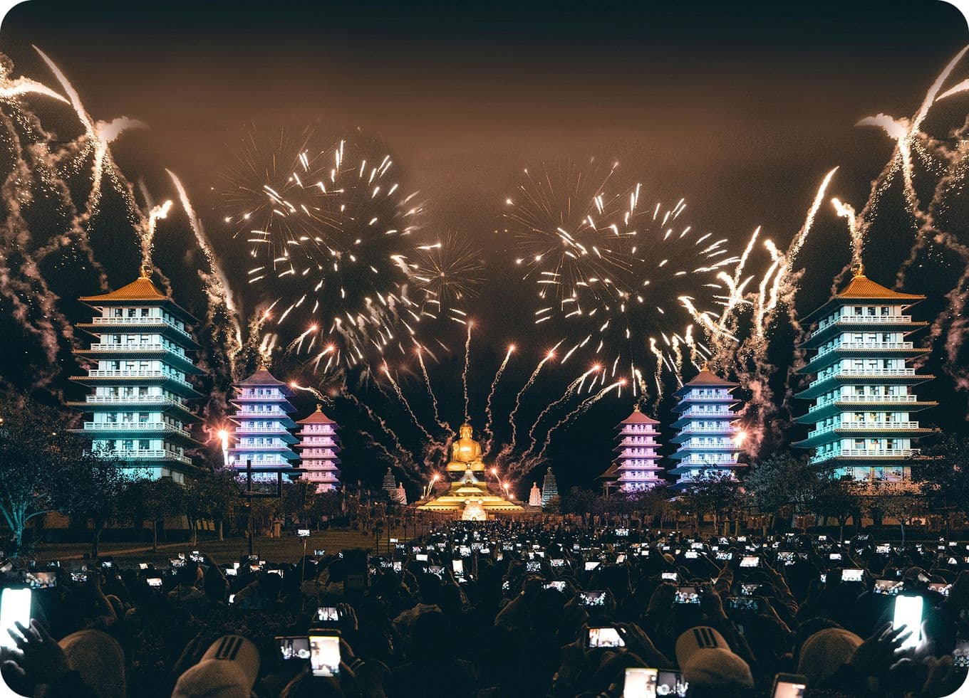 Fireworks illuminate the night sky above a large crowd, with pagodas and a golden statue in the background, as people capture the moment.