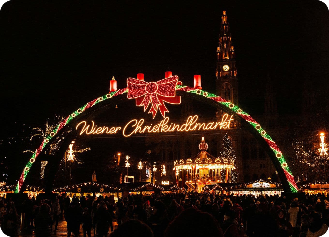 Illuminated entrance to Wiener Christkindlmarkt at night, with festive lights, a carousel, and bustling crowd.