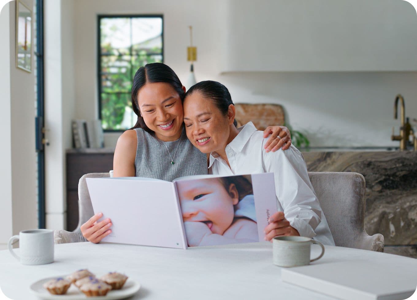 Two women sit at a table, smiling while looking at a photo album. Cups and pastries are on the table in a bright, modern room.
