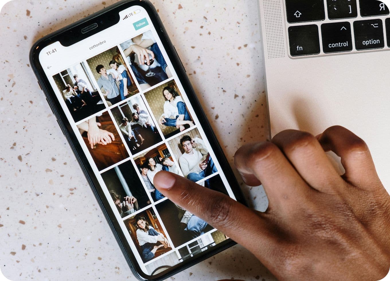 Person scrolling through a photo gallery on a smartphone next to a laptop on a speckled surface.