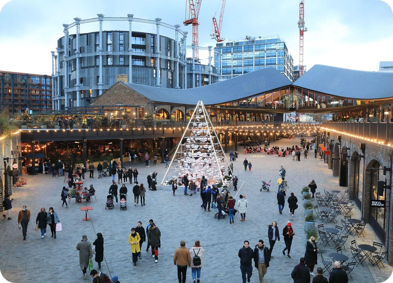 Busy urban plaza at dusk with illuminated triangular holiday tree installation, shoppers and surrounding shops.