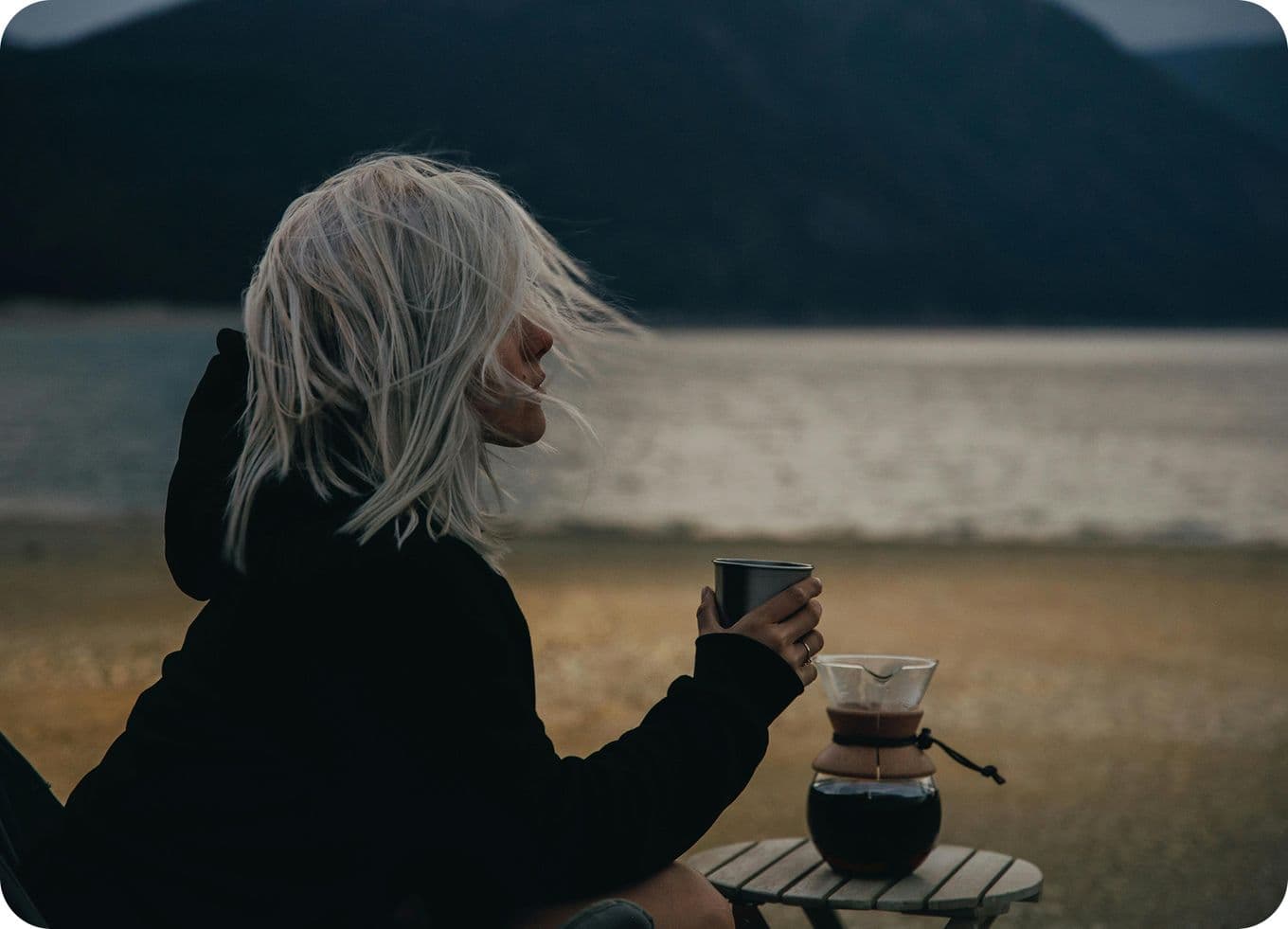Person with white hair sits by a lake, holding a cup, with a coffee pot nearby; mountains in the background.
