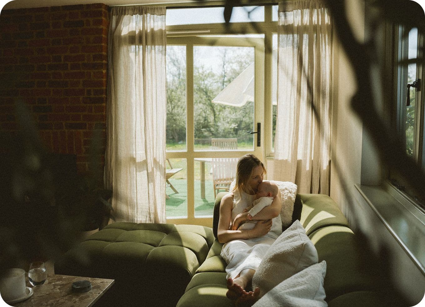 Woman sitting on a green sofa by a window, holding a baby wrapped in a white blanket, with sunlight streaming in through sheer curtains.