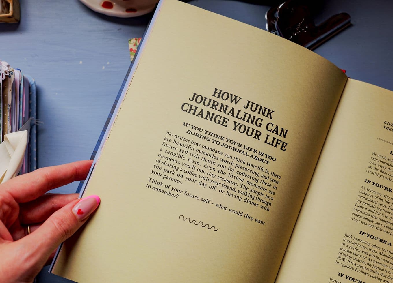 Hand with pink-painted nails holds open book titled "How Junk Journaling Can Change Your Life" on a blue desk.
