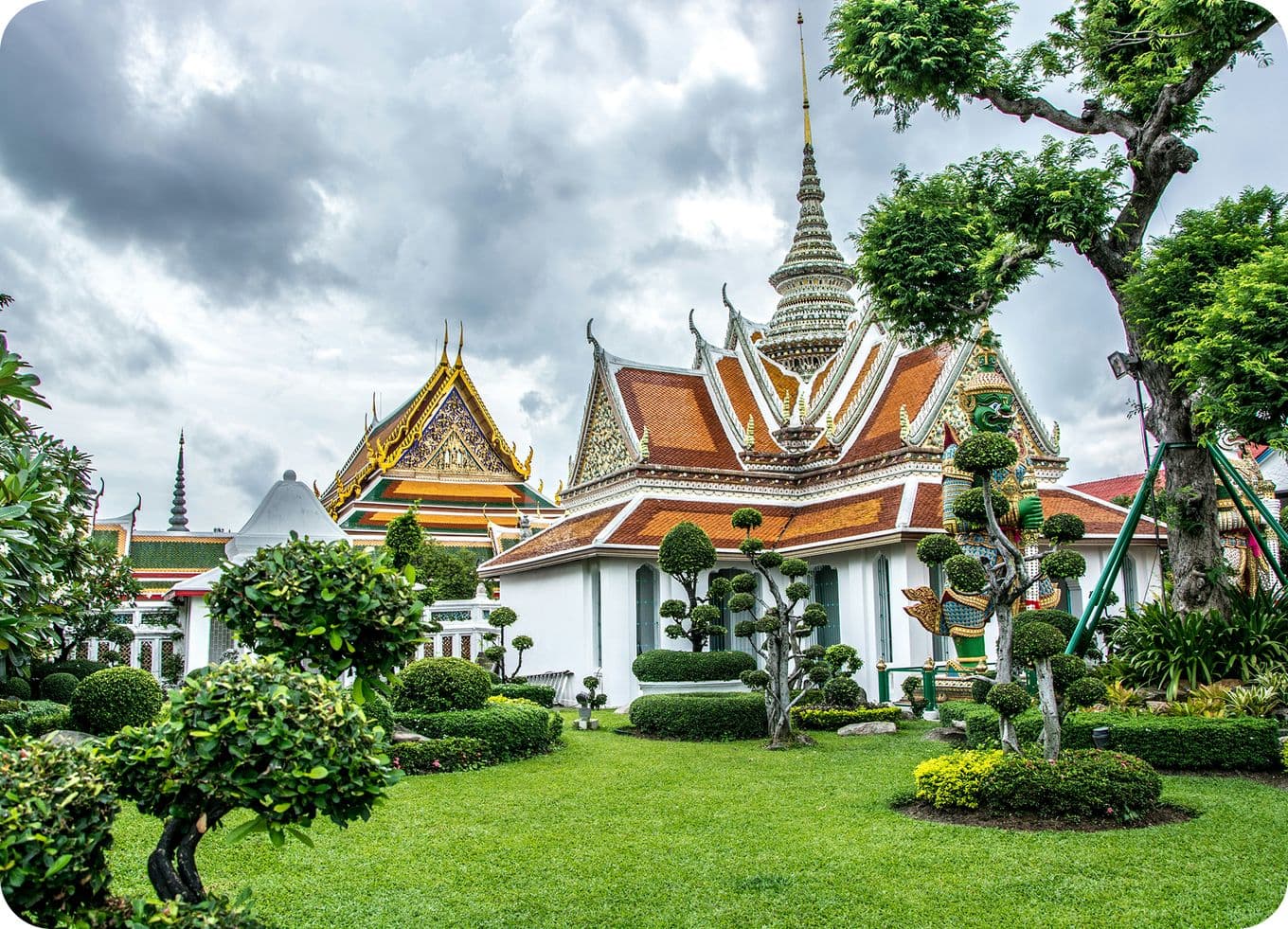 Ornate temple with intricate roofs and spires, surrounded by manicured gardens and topiary, under a cloudy sky.